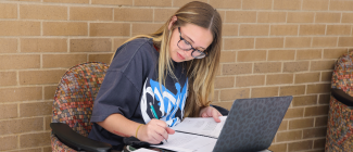 Female student taking notes on paper and her laptop while sitting at a desk